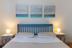 Ein Bett mit blauem Kopfteil, zwei Nachttischlampen und drei Gemälde vom Meer an der Wand.
