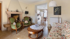 A living room with a fireplace, green armchairs, a round wooden table, a floral sofa, and a kitchen in the background.