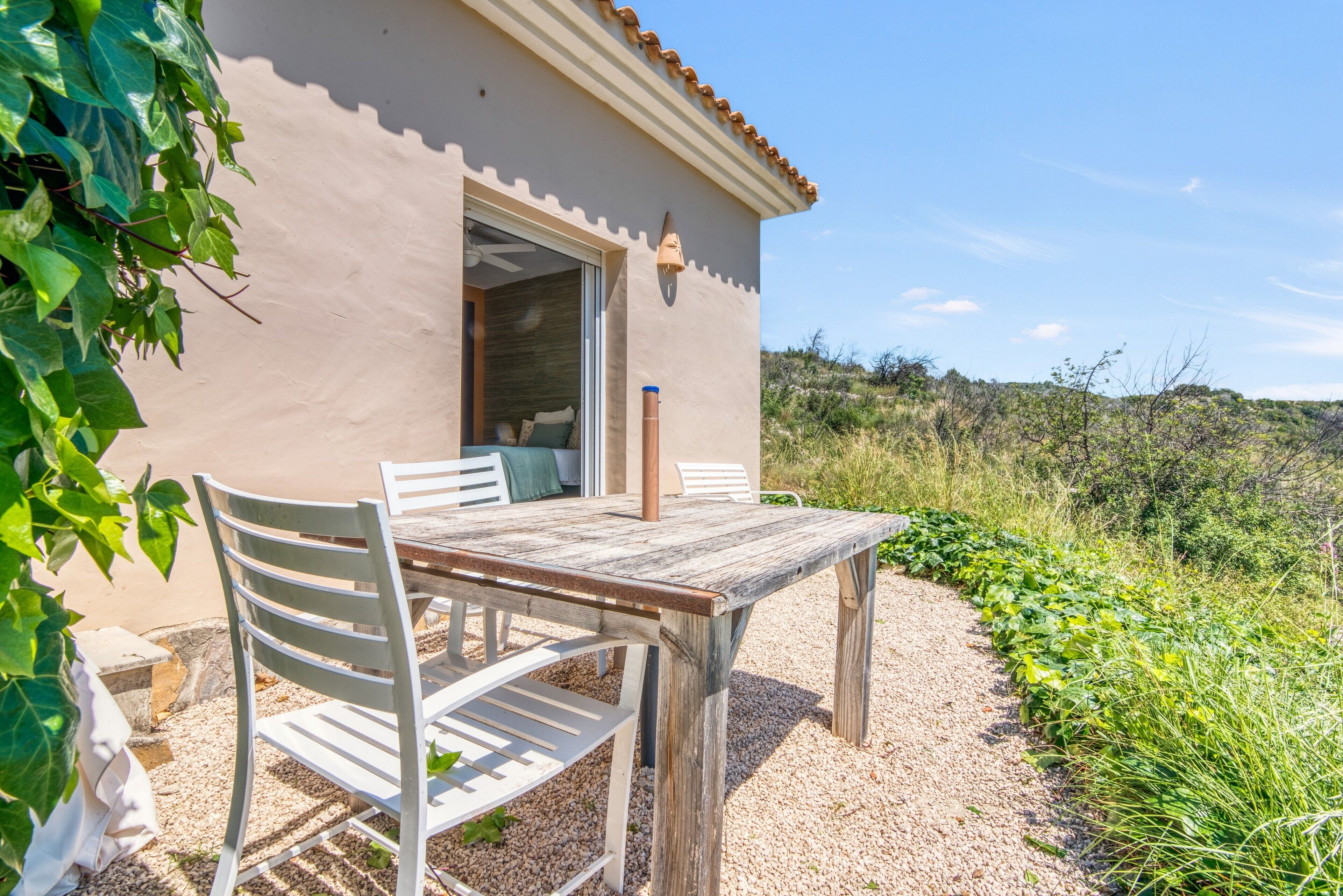 A rustic outdoor seating area with a table and chairs, a building entrance, and a gravel path leading to a natural landscape.