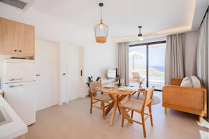 Een moderne keuken met witte kasten, een witte koelkast, een eethoek met een ronde tafel en rieten stoelen, en uitzicht op de zee door grote ramen.