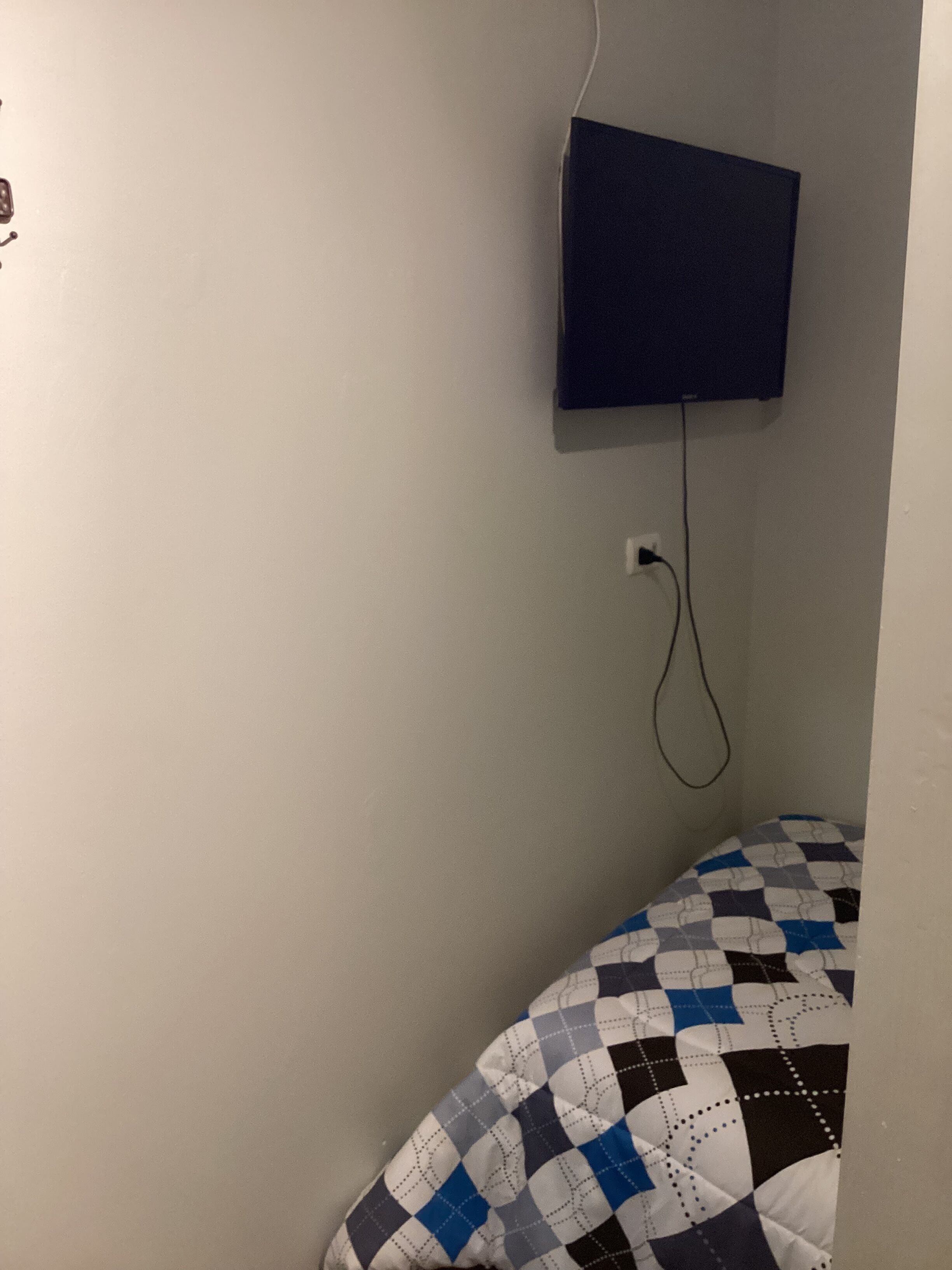 Una habitación pequeña con una cama individual, un televisor montado en la pared y un enchufe.