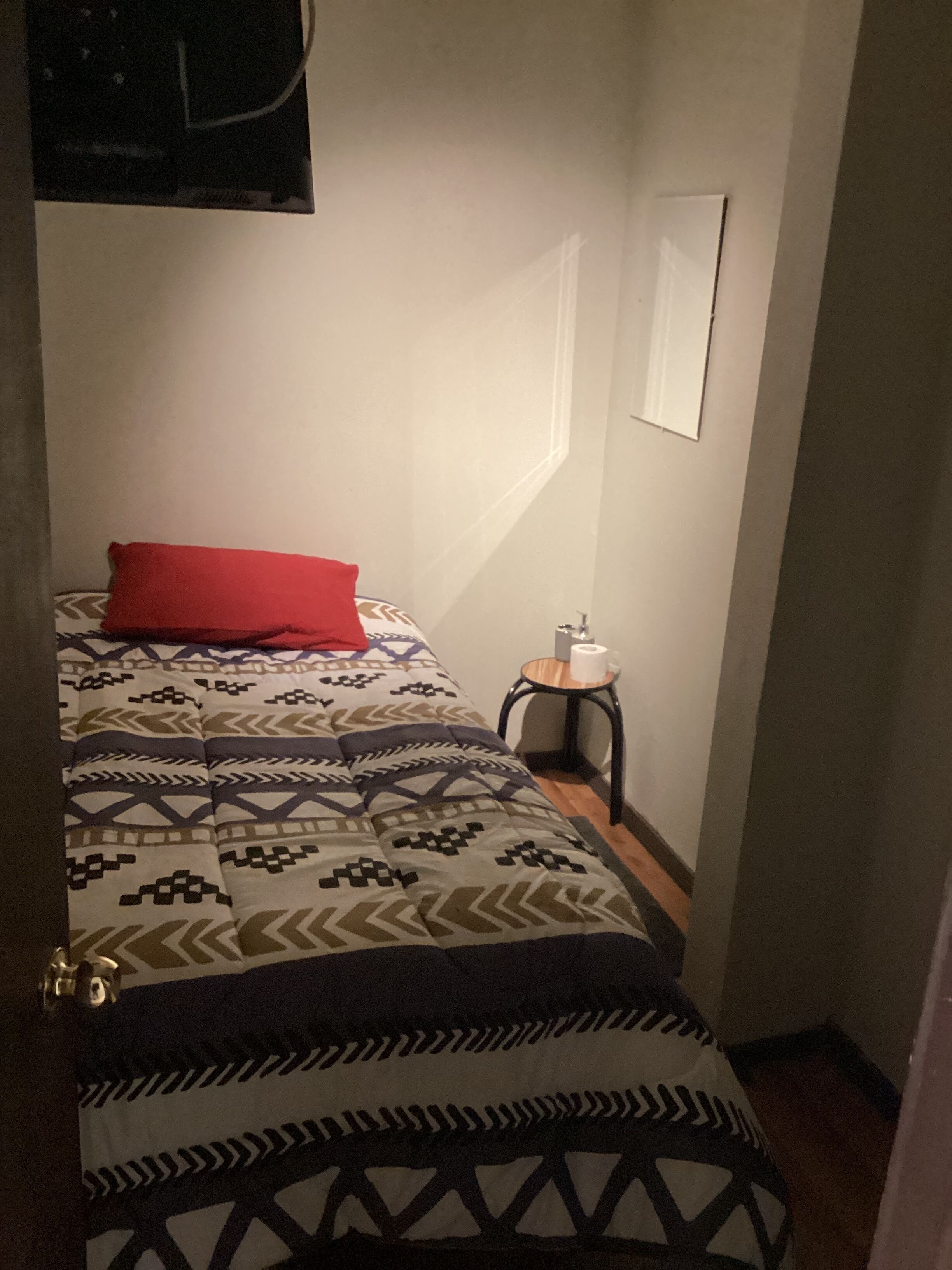 Una habitación pequeña y estrecha con una cama que tiene una colcha y almohadas estampadas, una mesita de noche con un rollo de papel higiénico, y un cuadro en la pared.