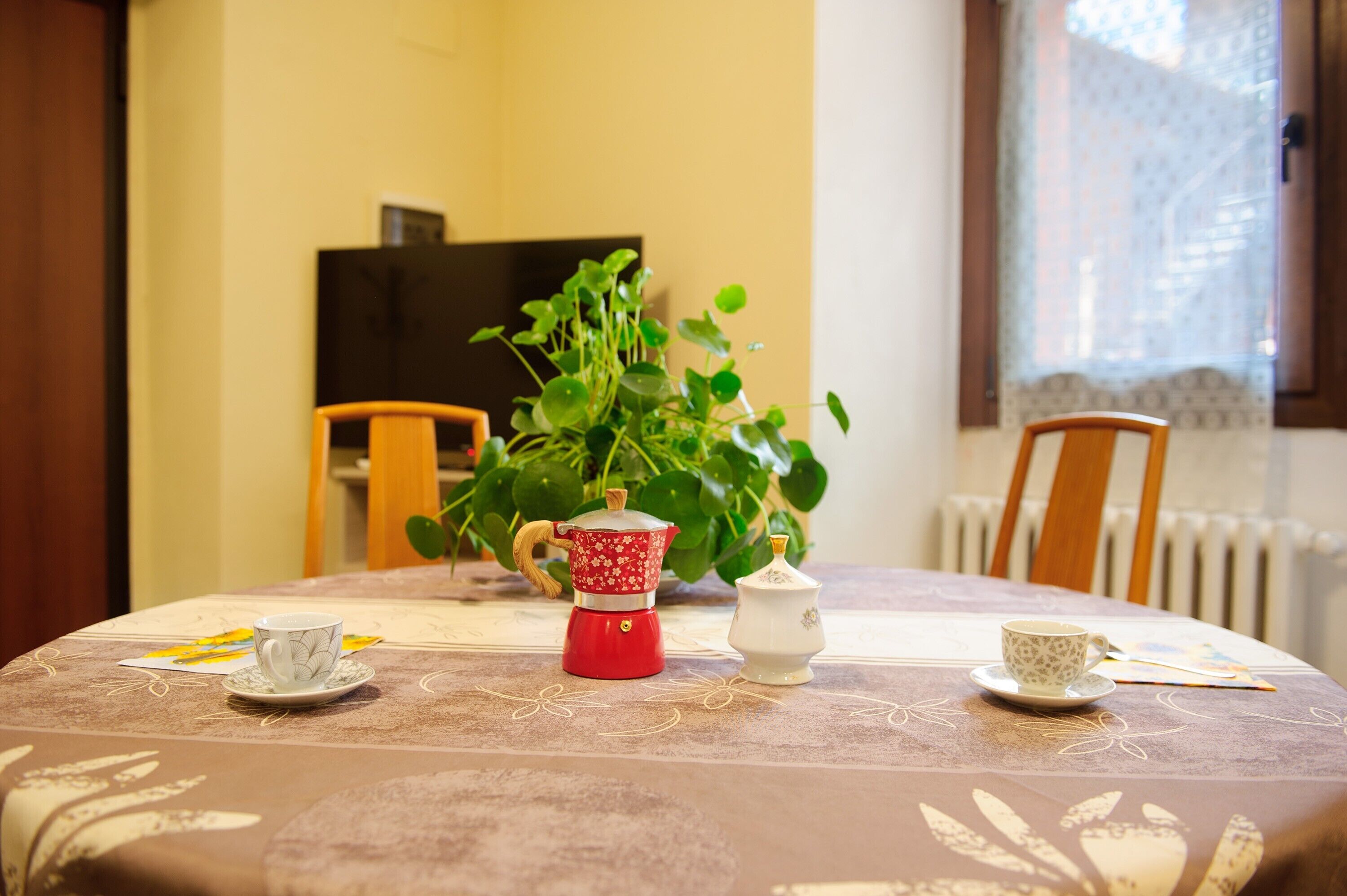 Une table de salle à manger ronde, recouverte d’une nappe à motifs floraux, une cafetière rouge, une théière blanche et deux tasses.