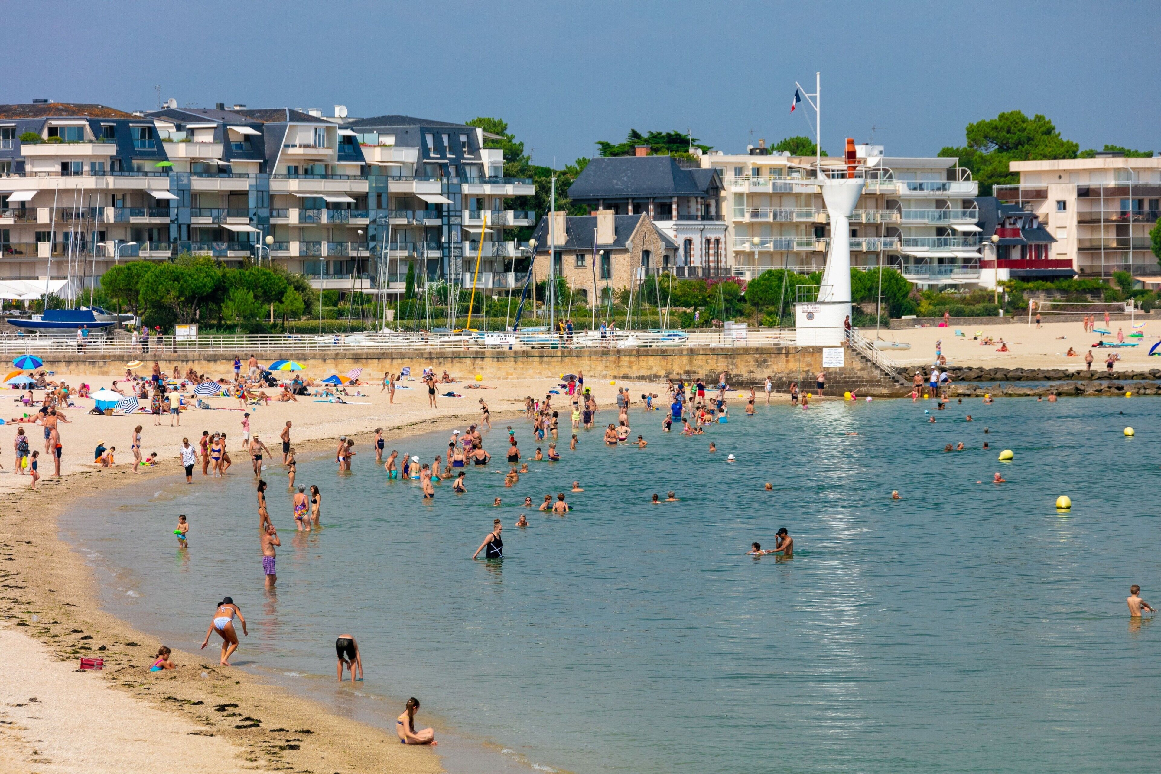 Ein Strand mit badenden und sonnenden Personen, einem Leuchtturm und modernen Gebäuden im Hintergrund.