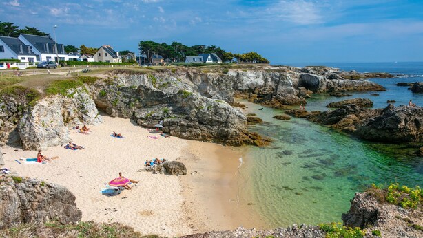 Une plage côtière avec des formations rocheuses, une eau claire et des maisons en arrière-plan.