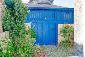 Une porte bleue, un jardin avec des fleurs rouges et un bâtiment doté d’un toit en pente.