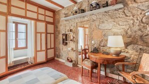 A traditional room with exposed stone walls, wooden beams, a bed, a table with a lamp, and a chair.