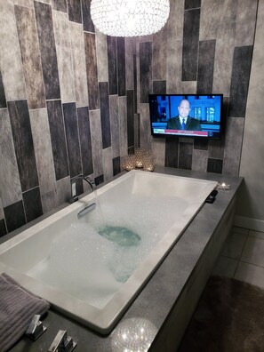 A modern bathroom with a large bathtub, a television mounted on the wall, and a decorative light fixture. - SolunaSpa Retreat Chefs Kitchen 10 Min to Beach (Long Beach)