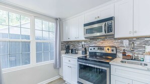 A modern kitchen with white cabinets, a stainless steel stove, a microwave, and a large window. - 148 Rockdale Street Tiny House (Myrtle Beach)