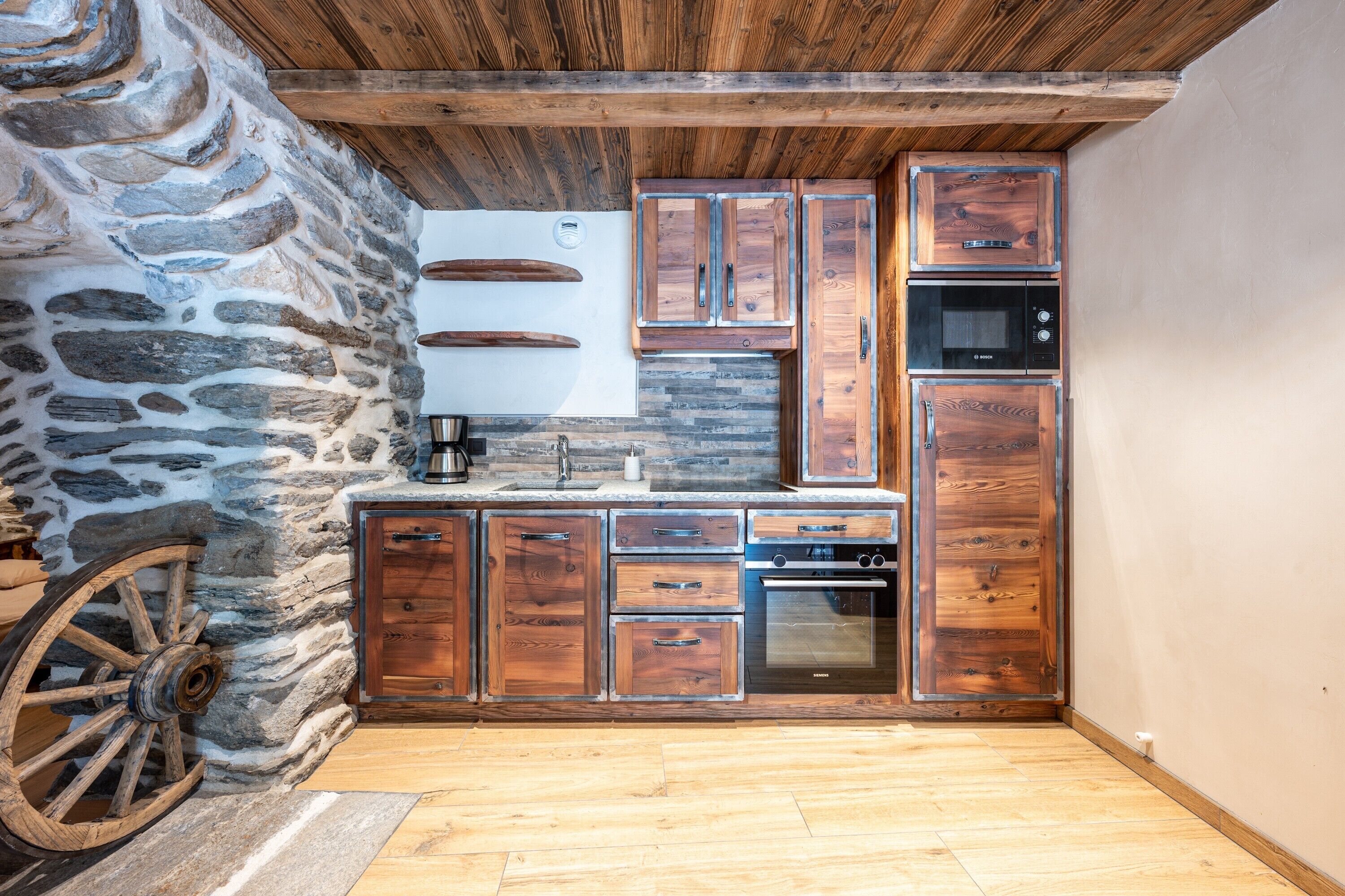 A rustic kitchen with wooden cabinets, a stone wall, and a wooden ceiling.
