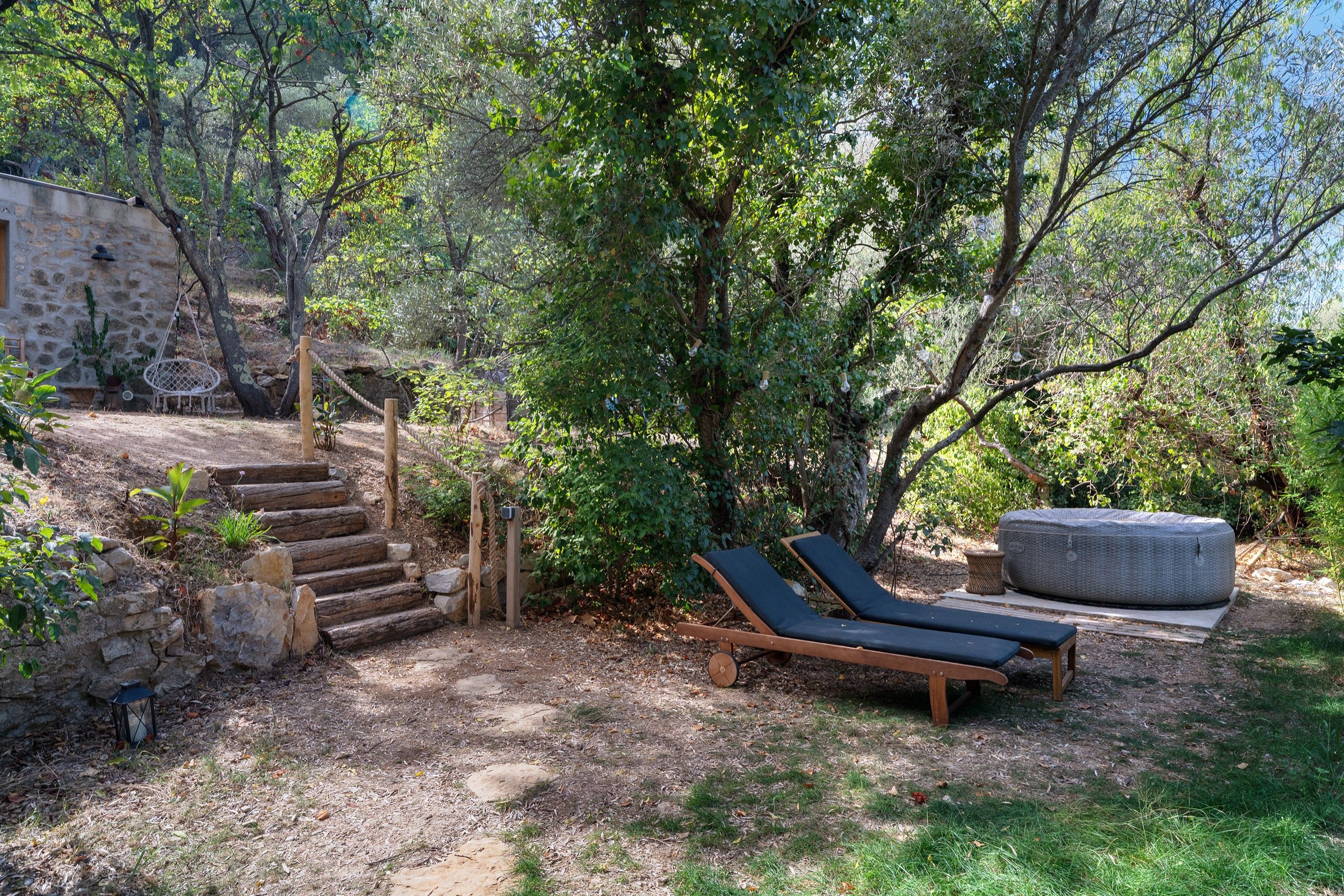 A rustic outdoor area with a stone wall, wooden steps, a hanging chair, and a lounge chair under a tree.