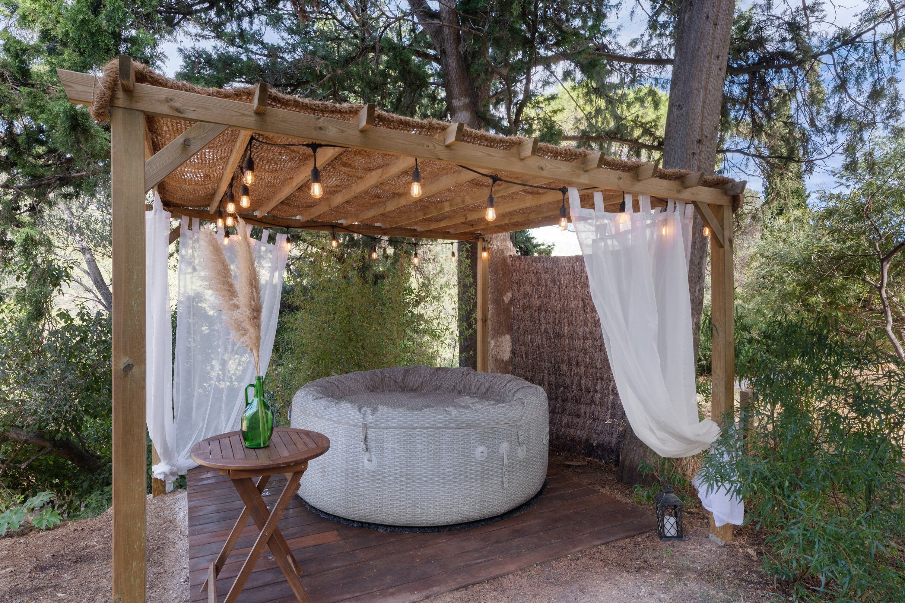 Une pergola en bois décorée de guirlandes lumineuses, un fauteuil rond en forme de sac de haricots et une petite table avec une bouteille.