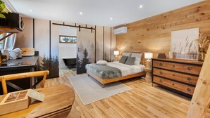 Un dormitorio moderno con piso de madera, una cama con cubrecama gris y blanco, un tocador de madera, un escritorio con una silla y un ventanal grande.