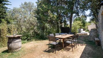 Un'area pranzo all'aperto con tavolo e sedie in legno, circondata da alberi e un muro di pietra.