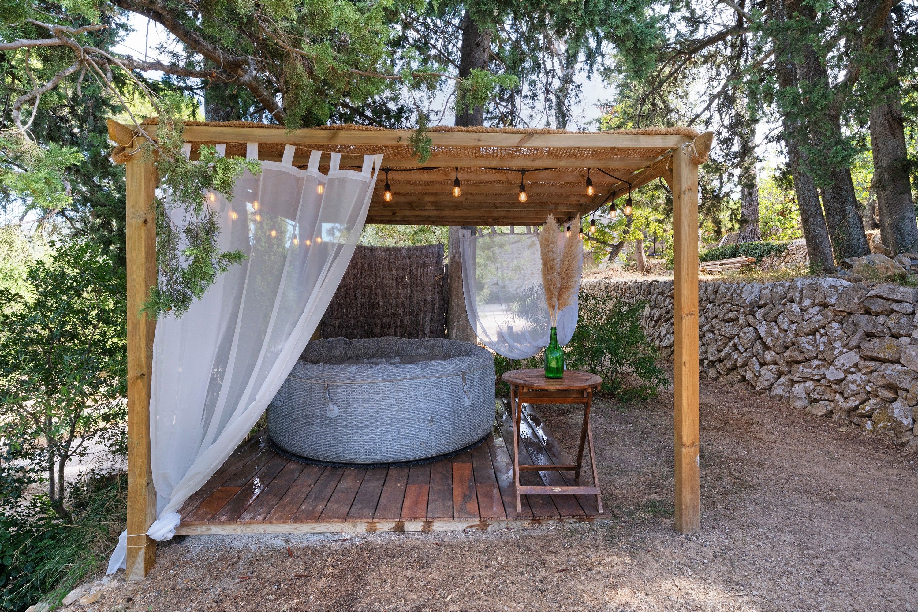 Uma pérgola de madeira com uma cama e uma pequena mesa, cercada por árvores e um muro de pedra.