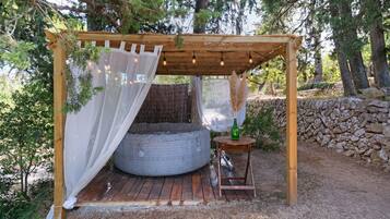 Uma pérgola de madeira com uma cama e uma pequena mesa, cercada por árvores e um muro de pedra.