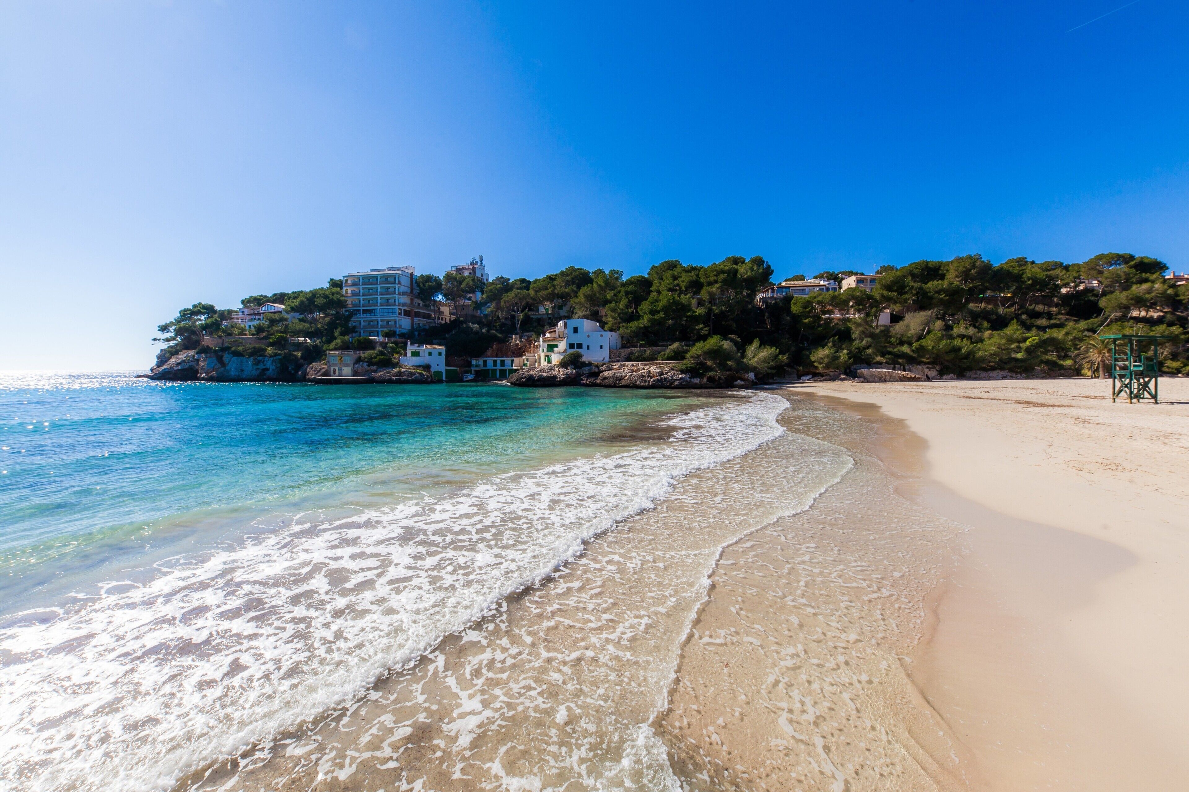 A beach with clear turquoise water, a sandy shore, and buildings on a hillside.