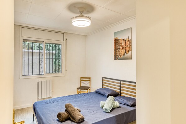 A bedroom with a bed, a chair, a radiator, a window with bars, and a cityscape painting.