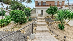 A two-story house with a stone staircase leading to a door, surrounded by a garden with various plants and a small tree.
