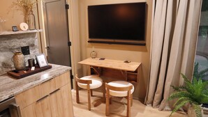 A compact kitchen with a marble countertop, a wooden dining table, two white cushioned chairs, a flat-screen TV mounted on the wall, and a potted plant.