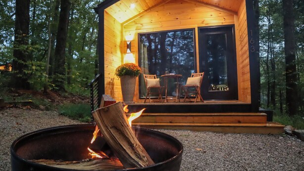 A cabin with a fire pit in the foreground, a wooden deck with two chairs and a table, and a large window with a view of the forest.