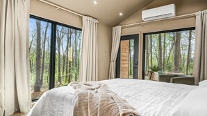 A bedroom with a large bed, a desk, and a view of a forest through the windows.