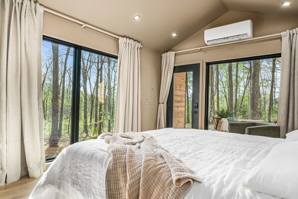 Un dormitorio con una cama grande, un escritorio y vistas a un bosque a través de las ventanas.