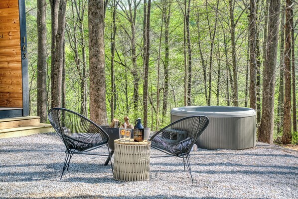 Un área de estar al aire libre con dos sillas, una mesa redonda pequeña y un jacuzzi, rodeada de un bosque.