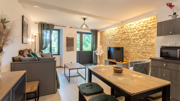 Une cuisine moderne avec un mur de pierres apparentes derrière le plan de travail, un coin repas avec une table en bois et des bancs, et un coin salon confortable avec des coussins.