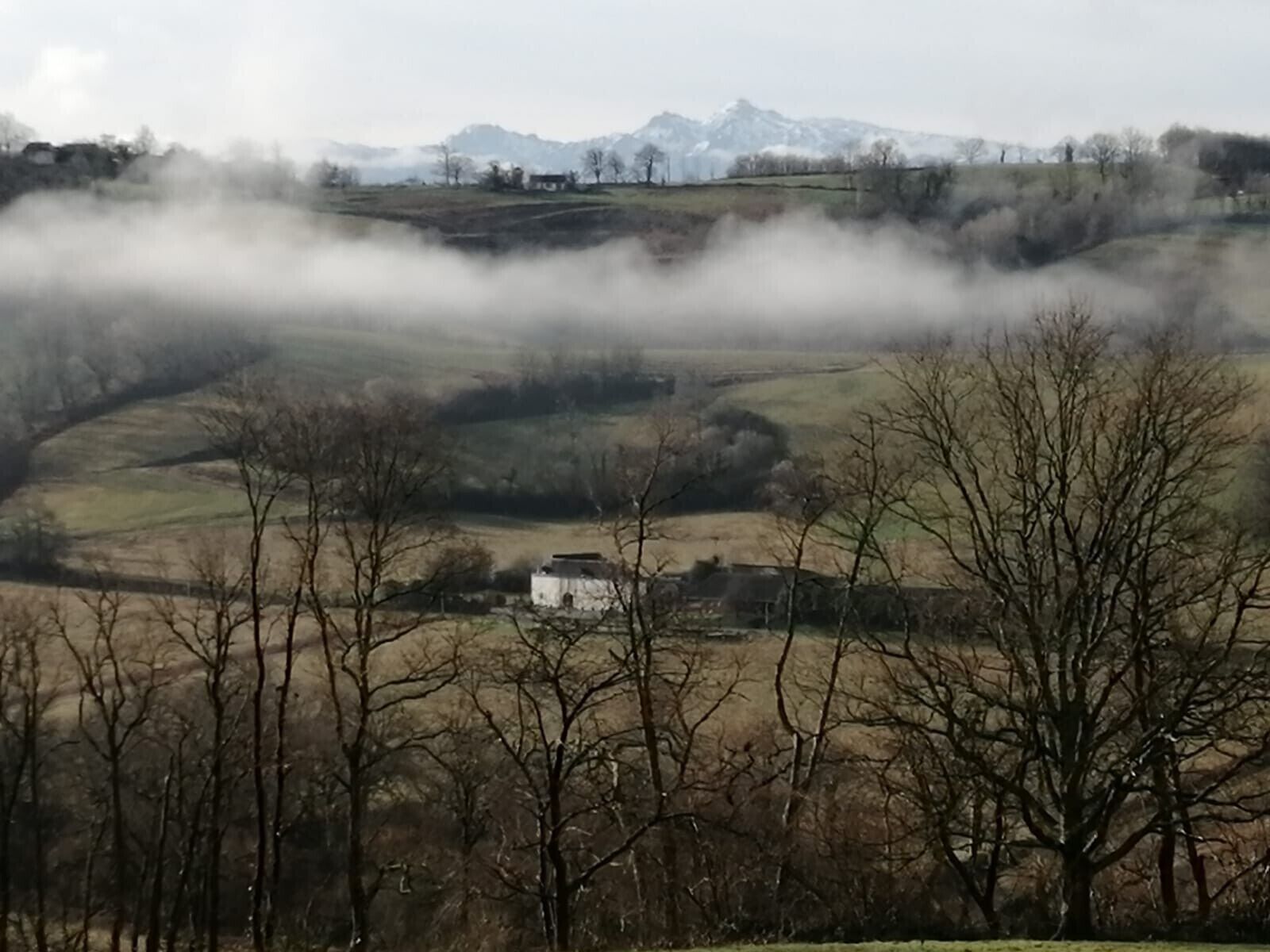 Un paysage rural avec des collines ondoyantes, un bâtiment blanc et une chaîne de montagnes à l’horizon.