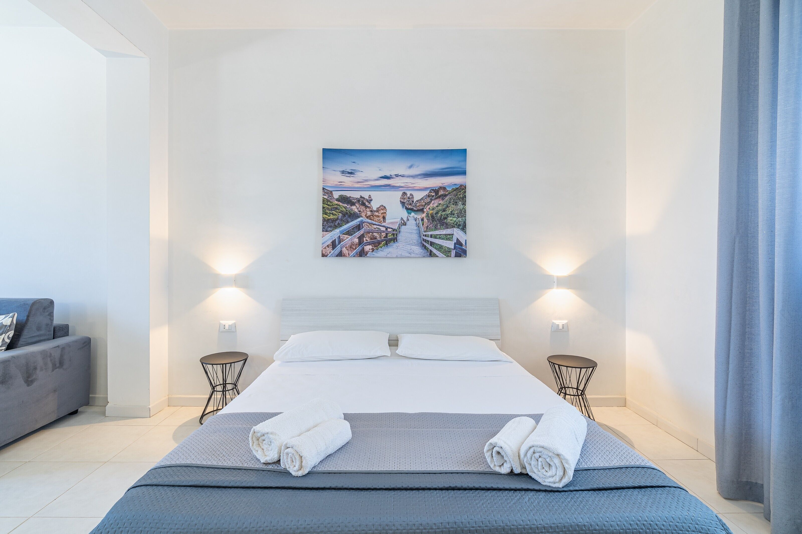 Ett modernt hotellrum med en stor säng, en tavla som föreställer ett kustlandskap, två sängbord och en grå soffa.