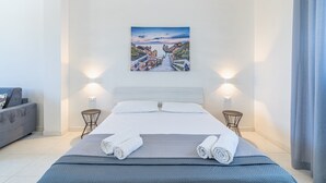 Une chambre d’hôtel moderne avec un grand lit, un tableau représentant un paysage côtier, deux tables de chevet et un canapé gris.