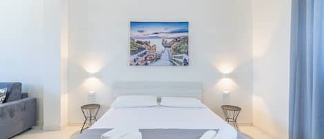 A modern hotel room with a large bed, a painting of a coastal landscape, two bedside tables, and a grey sofa.