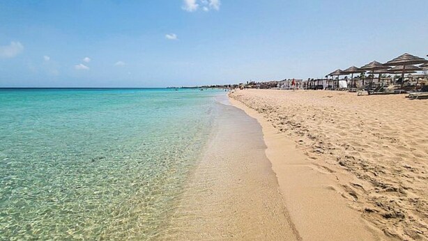 Una playa con agua turquesa cristalina, una orilla de arena y una hilera de construcciones frente al mar.