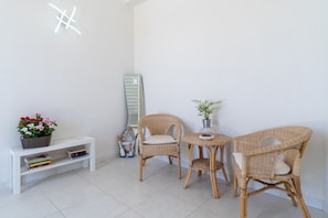 Una habitación con una consola blanca, dos sillas de mimbre, una pequeña mesa redonda con una planta en maceta y un espejo grande apoyado contra la pared.