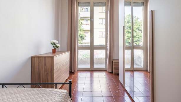 Une chambre à coucher comprenant un lit, une commode en bois, une plante en pot et une porte-fenêtre coulissante donnant sur un espace extérieur.