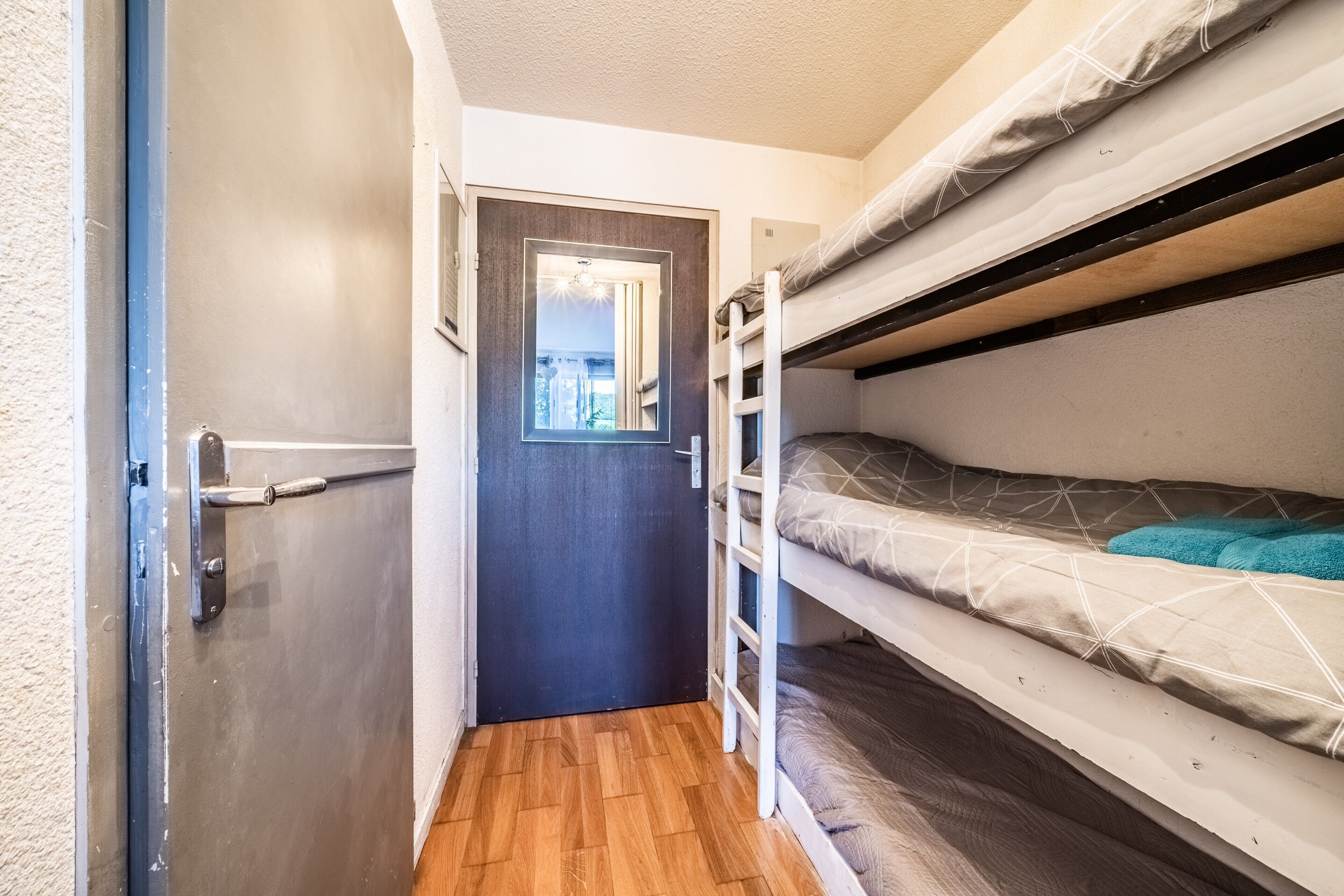 Une petite pièce avec un lit superposé, une porte dotée d’un miroir et un plancher en bois.