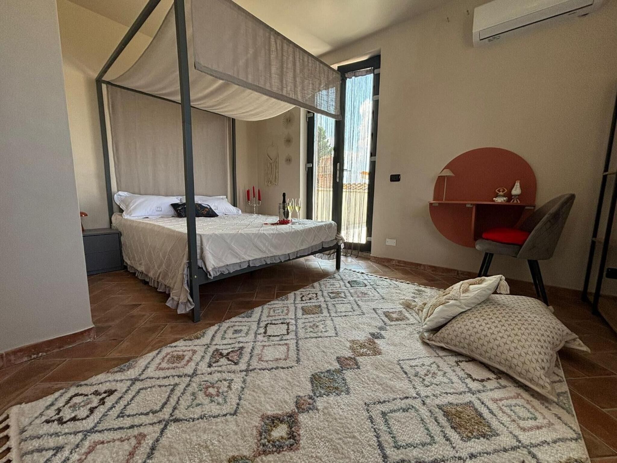 A four-poster bed with a canopy, a small desk with a chair, and a patterned rug on a tiled floor.