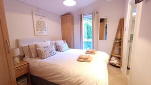 Une chambre à coucher comprenant un lit, des tables de chevet, une fenêtre avec des rideaux, une armoire en bois, un miroir et une œuvre d’art accrochée au mur.