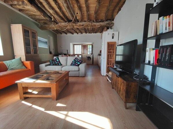 A living room with a sofa, coffee table, TV, bookshelf, and a kitchen area with a washing machine.