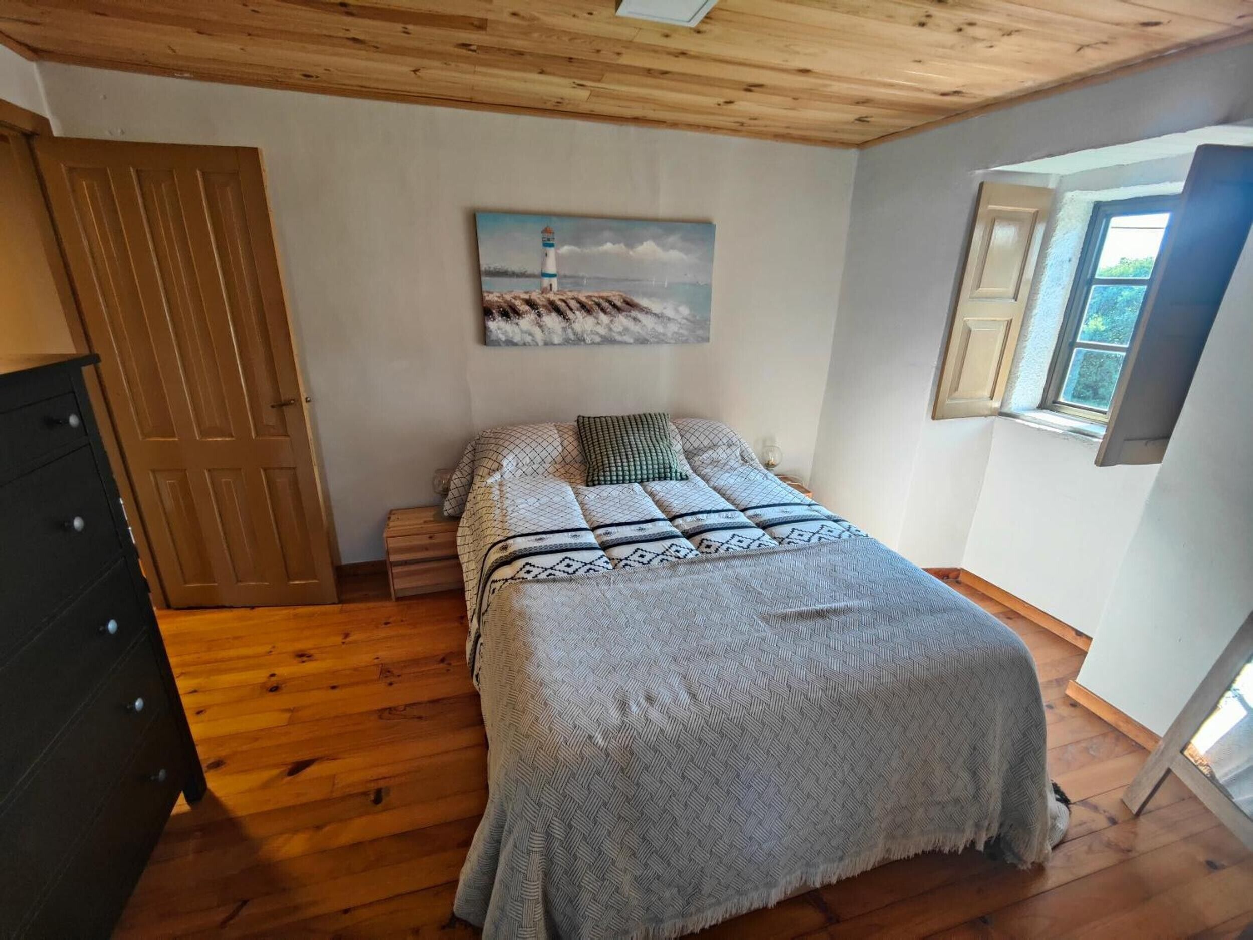 Um quarto com piso de madeira, uma cama com manta estampada, um quadro de um farol, cômoda de madeira e uma janela com vista.