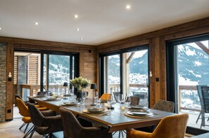 A dining area with a long table set for six, large windows with mountain views, wooden walls, and a stone accent wall. - Chalet "La Vora" with Mountain Views, Private Terrace and Wi-Fi (Châtel)