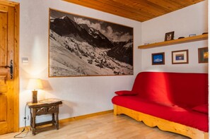 A room with a red sofa, a wooden table with a lamp, a large mountain landscape painting, and a wooden ceiling. - Apartment 'Appartement Super Chatel' with Mountain View, Balcony and Wi-Fi (Châtel)