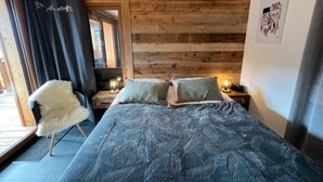 A bedroom with a wooden slat wall, a bed with patterned bedding, a fluffy chair, and a nightstand with a lamp. - Apartment 'Petite Grange 202' with Mountain View, Private Terrace and Wi-Fi (Unknown)