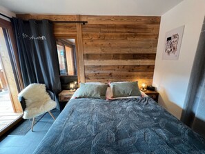 A bedroom with a wooden slat wall, a bed with patterned bedding, a fluffy chair, and a nightstand with a lamp.
