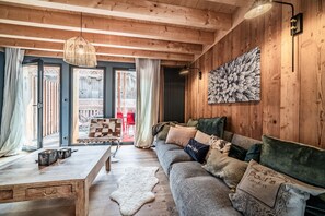 A cozy living room with a wooden ceiling, a sofa with various cushions, a coffee table, and a large window with curtains.