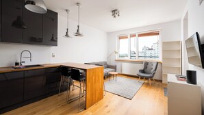 Une cuisine moderne avec des meubles noirs, un îlot central en bois et des tabourets de bar. Le salon comprend un canapé gris et un fauteuil, ainsi qu’une grande fenêtre donnant sur les bâtiments environnants.