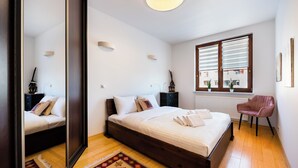 A bedroom with a large bed, a mirror, a window with blinds, and a chair.