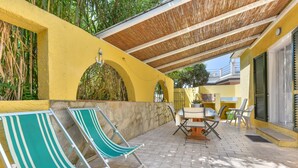 Un espace patio avec un mur jaune, une pergola, deux fauteuils de jardin à rayures, une table avec des chaises et un petit jardin.
