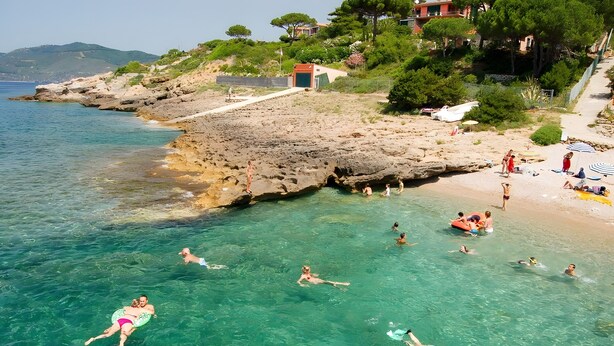 Une scène de plage côtière avec une eau claire, un rivage rocheux, et des gens qui nagent et prennent des bains de soleil.